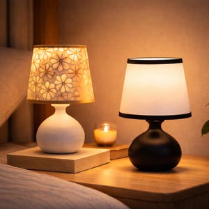 May include: Two small decorative lamps with shades. One lamp has a white base and a floral patterned shade. The other lamp has a black base and a white shade with a black trim. A lit candle sits between the lamps.