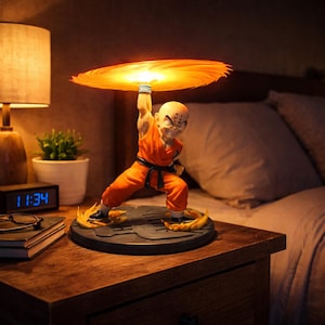 Op de afbeelding: Een lamp met een beeldje van Krillin uit Dragon Ball Z, in een oranje gi, die een gloeiende oranje energieschijf vasthoudt. De lamp staat op een donkerhouten nachtkastje, met een klok en een lamp op de achtergrond.