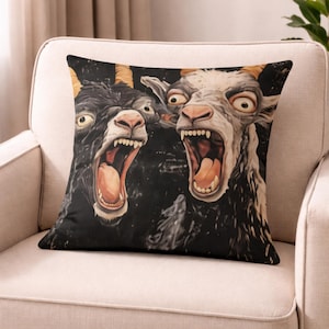 Può includere: Cuscino quadrato con un'immagine dipinta digitalmente di due capre con la bocca spalancata e espressioni esagerate. Le capre sono nere e bianche con corna marroni su uno sfondo scuro. Il cuscino è su una sedia color crema.