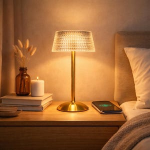 May include: A gold-colored table lamp with a textured lampshade sits on a wooden nightstand. A lit candle, a brown glass vase with dried flowers, and a smartphone are also on the nightstand. The lamp is next to a bed with a white pillow.