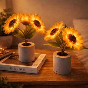 May include: Two illuminated artificial sunflower arrangements in grey concrete pots. The sunflowers have bright yellow petals and dark brown centres. The arrangements sit on a wooden surface, with a book titled "KINFOLK".