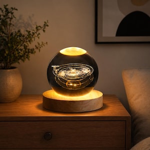 Op de afbeelding: Een bolvormig nachtlampje met een 3D-ontwerp van het zonnestelsel. De heldere glazen bol staat op een houten voet, die de planeten en hun banen verlicht. Het licht geeft een warme gloed, geschikt voor een slaapkamer of woonkamer.