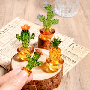 Op de afbeelding: Glazen cactus- en ananasbeeldjes met oranje en groene details. De cactusbeeldjes hebben kleine oranje en blauwe bloemaccenten. Het ananasbeeldje heeft een groene top. De beeldjes staan op een houten voet.
