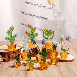 Puede incluir: Una colección de esculturas de cactus de vidrio de varias formas y tamaños, cada una en una pequeña maceta de color ámbar. Los cactus son verdes con detalles rojos, azules y amarillos. Un jarrón de cristal transparente y un papel con texto en el fondo.