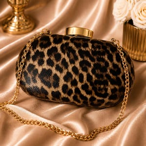 Peut inclure: Une pochette rigide avec un motif imprimé léopard marron et noir. Ce sac de forme ovale présente un fermoir métallique doré poli sur le dessus et une bandoulière en chaîne métallique dorée assortie fixée sur les côtés.