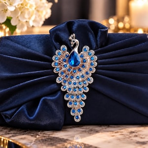May include: A navy blue velvet clutch bag featuring a large silver-toned peacock brooch centred on the front. The brooch is embellished with numerous small clear crystals and a prominent, teardrop-shaped blue gemstone at the peacock's body.