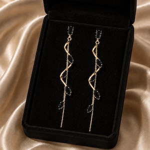 Könnte beinhalten: Ein Paar lange, goldfarbene Hängeohrringe mit einem filigranen Spiral-Drahtdesign, das mit kleinen, schwarzen, marquise-geschliffenen Steinen verziert ist. Jeder Ohrring hat ein gerades Kettenelement und einen Stecker, präsentiert in einer schwarzen Samtschatulle.