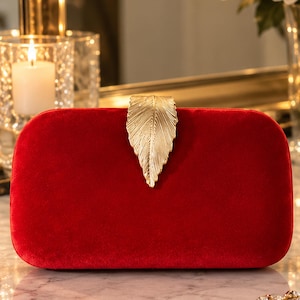 May include: A rectangular red velvet clutch bag featuring a prominent gold-toned metal clasp shaped like a textured leaf. The clutch has a smooth, soft-touch fabric exterior and a structured, rounded-edge design with a central metallic closure.