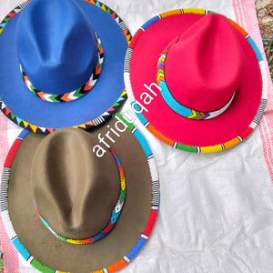 May include: Three felt hats in blue, red, and brown, each with a beaded band in a colourful geometric pattern. The hats have a classic fedora shape and are displayed on a white surface. The text "afriduqah" is visible on the image.