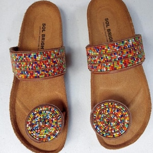 May include: A pair of brown leather sandals with colorful beaded accents. The sandals have a toe loop and a strap across the top of the foot, both adorned with small, vibrant beads. The footbed is a light brown color, and the brand name "SOL BRIGHT" is printed on the footbed.