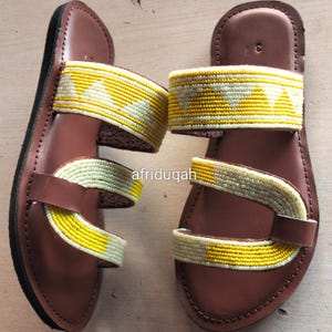 Puede incluir: Un par de sandalias de cuero marrón con correas de cuentas amarillas y blancas. Las sandalias tienen una suela plana y dos correas en la parte superior del pie. El texto "afriduqah" es visible en el lateral.