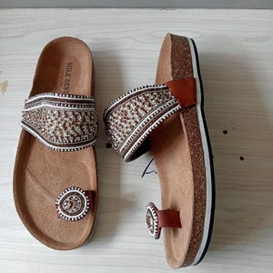May include: Brown sandals with beaded straps and toe loops. The sandals have a cork footbed and a white and black sole. The straps are decorated with a pattern of white, brown, and tan beads. The toe loop also has a beaded design.