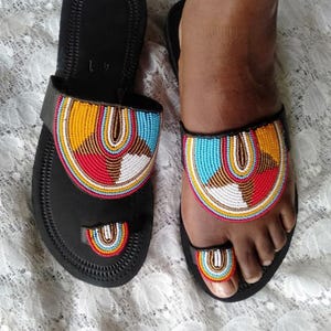 May include: Black sandals with colorful beaded designs. The sandals feature a toe strap and a wide strap across the top of the foot, both adorned with intricate patterns in red, yellow, blue, and white beads. The sandals are displayed on a white lace surface.