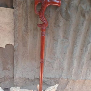 Op de afbeelding: Een handgemaakte houten wandelstok met een roodbruine afwerking. De handgreep heeft een olifantenkopontwerp en een gebogen grip. De lange, slanke schacht heeft gesneden details. Een decoratief en functioneel accessoire.