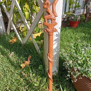 Op de afbeelding: Een houten wandelstok met ingewikkelde gravures. De stok heeft een figuur die bovenop een decoratieve kolom zit, met een gebogen handvat. Een gedetailleerde slang windt zich om de schacht. De stok is gemaakt van lichtbruin hout, ongeveer 150 cm hoog.