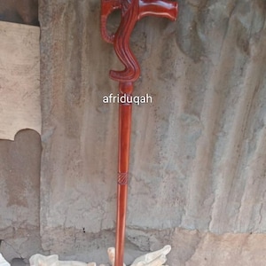 May include: A handcrafted wooden walking cane with a reddish-brown finish. The handle is carved in an elephant head design, with a curved support. The shaft is smooth with a decorative band near the top. The text "afriduqah" is visible.