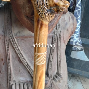 May include: Wooden walking cane with a carved lion's head handle. The cane is light brown with a natural wood grain and a decorative carved pattern near the top. The text "afriduqah" is printed on the cane.