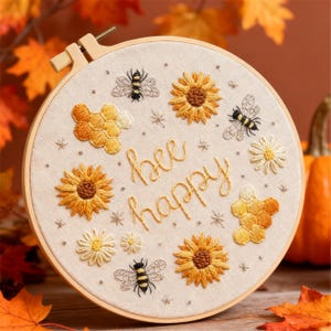 Puede incluir: Arte bordado en bastidor con girasoles, abejas y diseños de panal. La frase "bee happy" está bordada en cursiva. La obra está enmarcada en un aro de madera y rodeada de hojas de otoño.