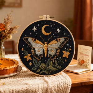 May include: A circular embroidery hoop with a black background, showcasing a butterfly design in gold, white, and grey thread. The design includes a crescent moon, stars, and floral elements. The hoop is displayed on a neutral surface.