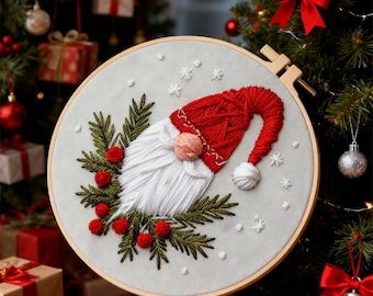 Könnte beinhalten: Gestickter Weihnachtszwerg in einem Holzreifen. Der Zwerg hat eine rote Mütze, einen weißen Bart und eine rosa Nase, umgeben von grünen Tannennadeln und roten Beeren. Der Hintergrund ist ein hellgrauer Stoff mit weißen Schneeflocken.