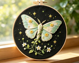 May include: A circular embroidery hoop containing black fabric with a hand-stitched moth featuring pale green and white wings. The moth is surrounded by yellow embroidered stars and small white flowers with green leaves on a vine extending downward.