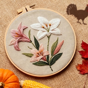 May include: Embroidered floral artwork in a wooden hoop. The design features pink and white lilies with green leaves. The background is a neutral linen fabric. A small pumpkin, corn, and a red leaf are in the foreground.