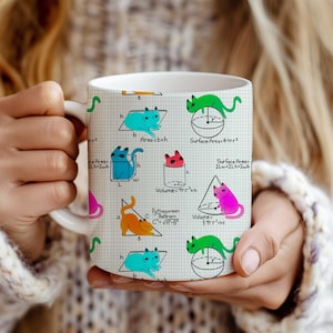May include: White ceramic mug featuring colorful cartoon cats illustrating geometric formulas. The cats are depicted in various poses, with formulas for area, volume, and the Pythagorean theorem. The mug is held by a person wearing a white sweater.