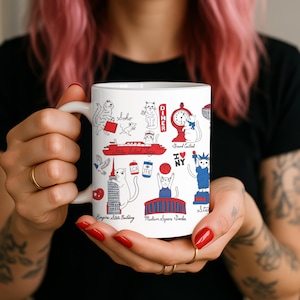 May include: White ceramic mug featuring illustrations of New York City landmarks and cats. The design includes the Empire State Building, the Statue of Liberty, and the words "Soho" and "Diner". The mug is held by a person with red nail polish.
