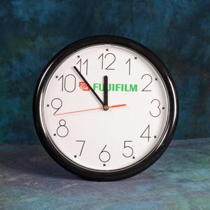 May include: A round wall clock with a black frame and a white face. The clock has black numerals, a red second hand, and the Fujifilm logo. The time is approximately 11:59.