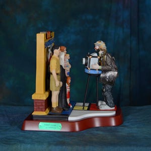 May include: A decorative figurine depicting a clown photographer taking pictures of a group of people. The figurine has a red base and a sign that says "FUNNY CLOWN". The clown is wearing a hat and a jacket.