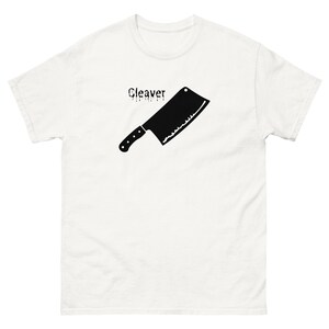 May include: White t-shirt featuring a black cleaver graphic with the word "Cleaver" above it. The cleaver is a kitchen tool with a rectangular blade and a handle with black dots. The shirt is made of a soft material.