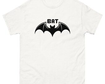 May include: White t-shirt featuring a black bat graphic with the word "BAT" above the bat's wings. The bat graphic is centered on the front of the shirt. The shirt has short sleeves and a crew neck.