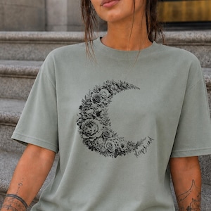 以下が含まれることがあります： セージグリーンの半袖Tシャツ。様々な花や葉で構成された三日月の黒いグラフィックプリントが施されています。月のグラフィックの下部には、筆記体で「sleepyhead」という文字がデザインされています。