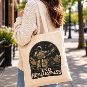 May include: A beige canvas tote bag featuring a graphic illustration of an astronaut standing on a lunar surface, holding a sign that reads TO A BETTER TOMORROW. Below the astronaut, the text END HOMELESSNESS is printed in bold lettering.