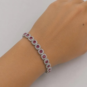 May include: A silver bracelet with square-cut, deep red gemstones, each encircled by smaller, clear stones. The bracelet is shown on a wrist, highlighting its design and sparkle. The bracelet is a piece of jewellery.