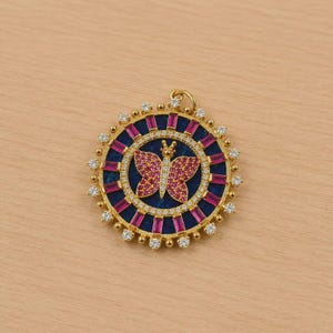 May include: A gold-toned pendant with a butterfly design. The pendant features a central butterfly shape with pink accents, surrounded by a blue and pink circular pattern. Small, clear stones and a gold loop complete the design.