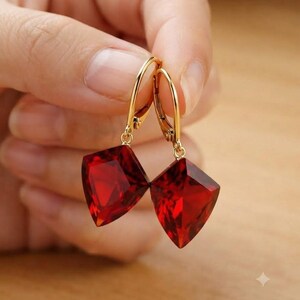 May include: A pair of gold leverback earrings featuring faceted, kite-shaped deep red gemstones. The earrings have a polished gold metal finish and a geometric, angular cut that catches the light, suspended from secure, hinged gold ear wires.