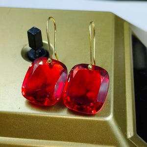 May include: A pair of red, square-cut gemstone earrings with gold-coloured hooks. The earrings are a vibrant red colour and appear to be faceted, catching the light. The gold hooks are simple and elegant.