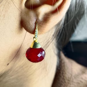 Puede incluir: Un pendiente dorado con una piedra roja facetada en forma de lágrima. El pendiente tiene una pequeña tapa dorada y un gancho adornado con pequeñas piedras transparentes. El pendiente se muestra sobre un fondo borroso.