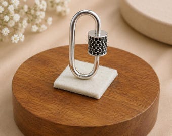 May include: A silver-toned oval carabiner-style jewelry clasp featuring a cylindrical section encrusted with small, dark black stones. The clasp has a smooth, polished metallic finish and a functional spring-loaded gate mechanism for securing items.