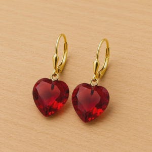 Puede incluir: Un par de pendientes dorados con cierre de palanca y gemas rojas facetadas en forma de corazón. Los pendientes se muestran sobre una superficie de madera marrón claro. Las gemas en forma de corazón son de un color rojo intenso.