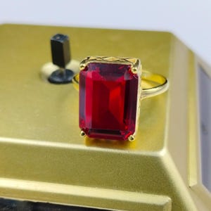 Może przedstawiać: Złoty pierścionek z dużym, prostokątnym, głęboko czerwonym kamieniem szlachetnym. Oprawa i oprawa pierścionka są w kolorze złotym, a kamień szlachetny jest punktem centralnym. Pierścionek jest osadzony na złotej powierzchni.