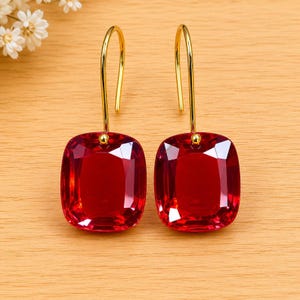 May include: A pair of red, square-cut gemstone earrings with gold-coloured hooks. The earrings are a vibrant red colour and have a faceted surface, reflecting light. The hooks are a simple, elegant design.