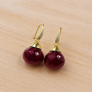 Puede incluir: Un par de pendientes colgantes dorados con esferas de piedras preciosas rojo oscuro facetadas. Los pendientes tienen una tapa dorada y un gancho con pequeñas piedras transparentes. Se muestran sobre una superficie de madera marrón claro.