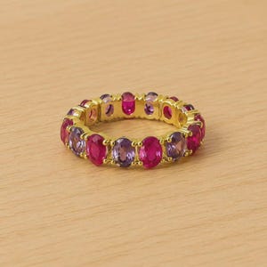 May include: A gold-toned ring featuring alternating oval-cut gemstones in shades of pink and purple. The ring is set on a light-colored wooden surface, showcasing the vibrant colors of the stones.