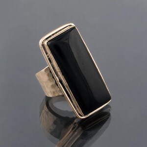Peut inclure: Une bague ornée d'une grande pierre noire rectangulaire polie, sertie dans une monture en métal doré. L'anneau de la bague présente une texture martelée distincte, contrastant avec la surface lisse et réfléchissante de la pierre noire.