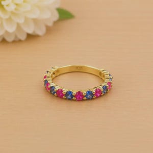 May include: A gold-colored ring with alternating pink and blue gemstones. The ring is set with small, round stones and has the number "925" engraved on the band. The ring is on a light brown surface.