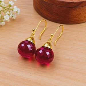 May include: A pair of gold-colored drop earrings with faceted, round, ruby-red pendants. The earrings are displayed on a light-colored wooden surface, with a wooden box in the background. Delicate white flowers are visible in the upper left corner.
