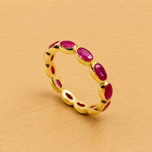 May include: A gold ring featuring a series of oval-cut, vibrant red gemstones. The gemstones are set in a continuous pattern around the ring's band. The ring is displayed on a light brown surface.