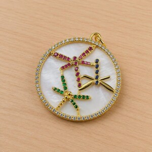 May include: Gold-tone pendant with a white center and three dragonfly designs. The dragonflies are adorned with red, green, and blue gemstones. The pendant is encircled by a row of clear gemstones, creating a sparkling effect.